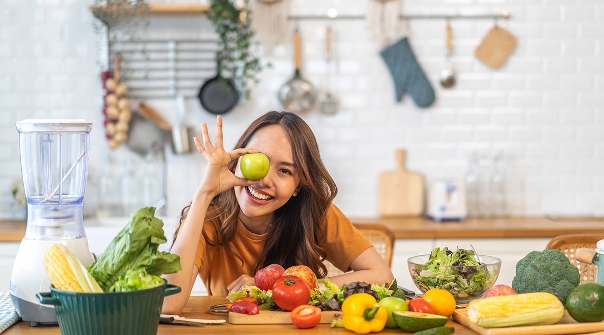 Junge, lachende Frau steht vor einem Küchentresen mit verschiedenem Gemüse und hält einen grünen Apfel vor ihrem Auge