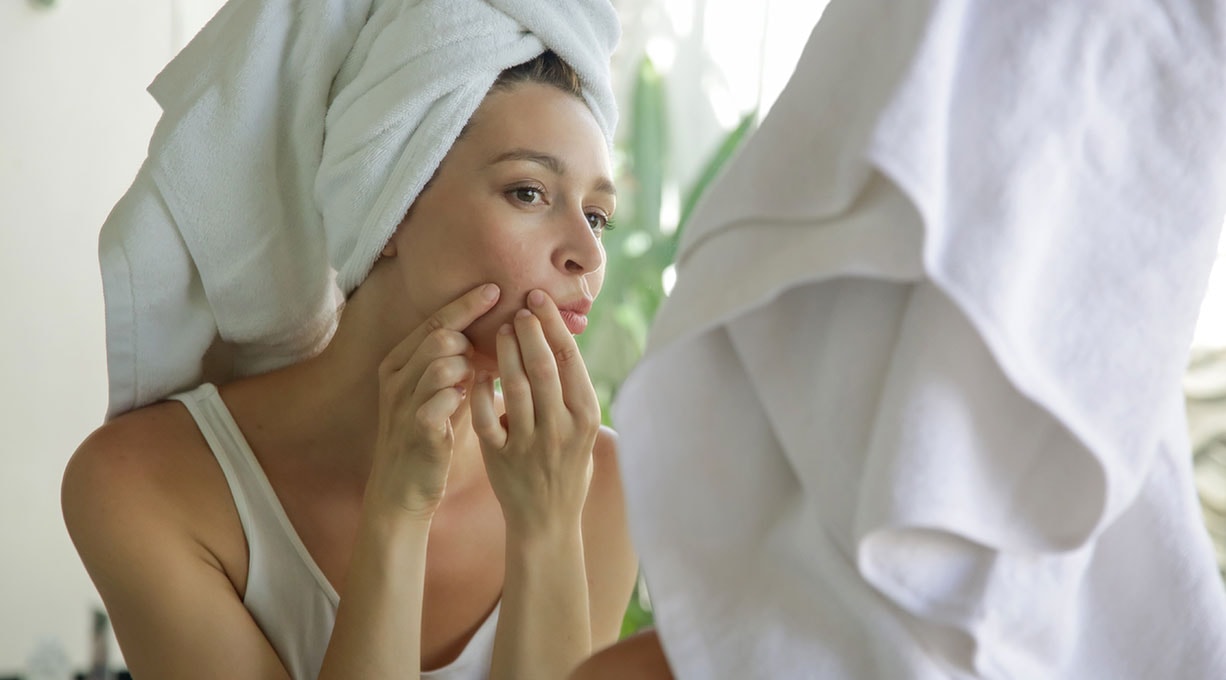 Eine Frau mit Handtuch berührt vor dem Spiegel die Gesichtshaut zur Hautpflege
