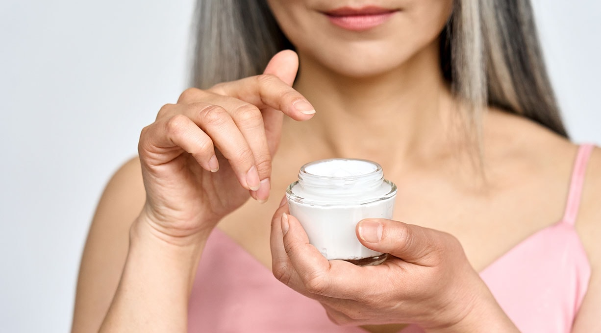 Frau hält offenen Cremetopf gegen Spätakne der Zeigefinger schwebt über der Creme