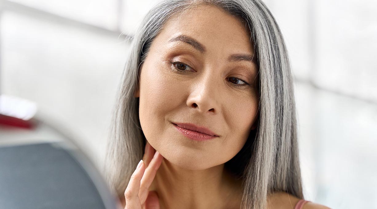 Frau mit langen grauen Haaren blickt in den Spiegel und berührt die Wange Fokus auf Hautbild
