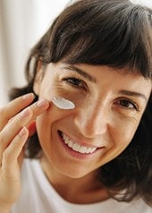 Eine breit lächelnde Frau trägt ein Hautpflegeprodukt auf ihr Gesicht auf.