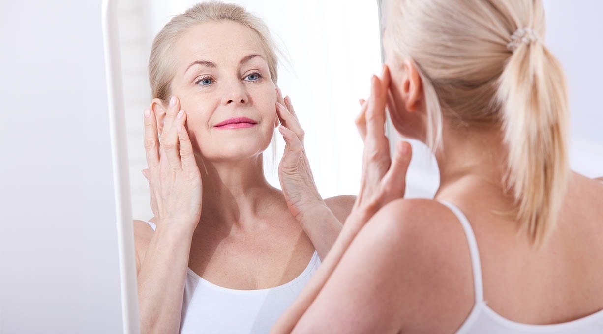 Ältere Frau mit blondem Pferdeschwanz berührt ihr Gesicht und betrachtet sich lächelnd im Spiegel