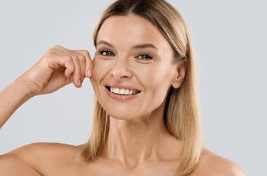 Frau mit blonden Haaren zieht sanft an ihrer Wange und lächelt in die Kamera