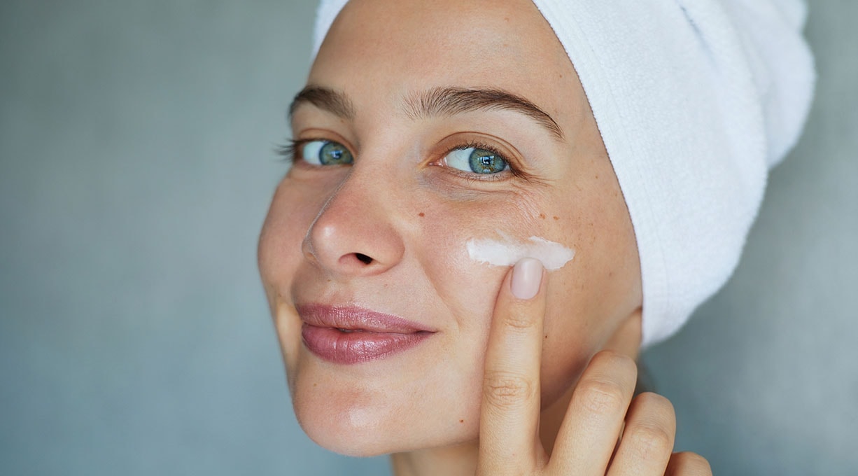 Frau mit Handtuch auf dem Kopf trägt Creme auf ihr Gesicht auf und lächelt