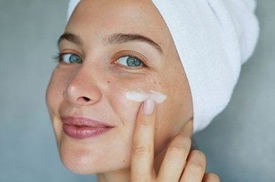 Frau mit Handtuch auf dem Kopf trägt Creme auf ihr Gesicht auf und lächelt