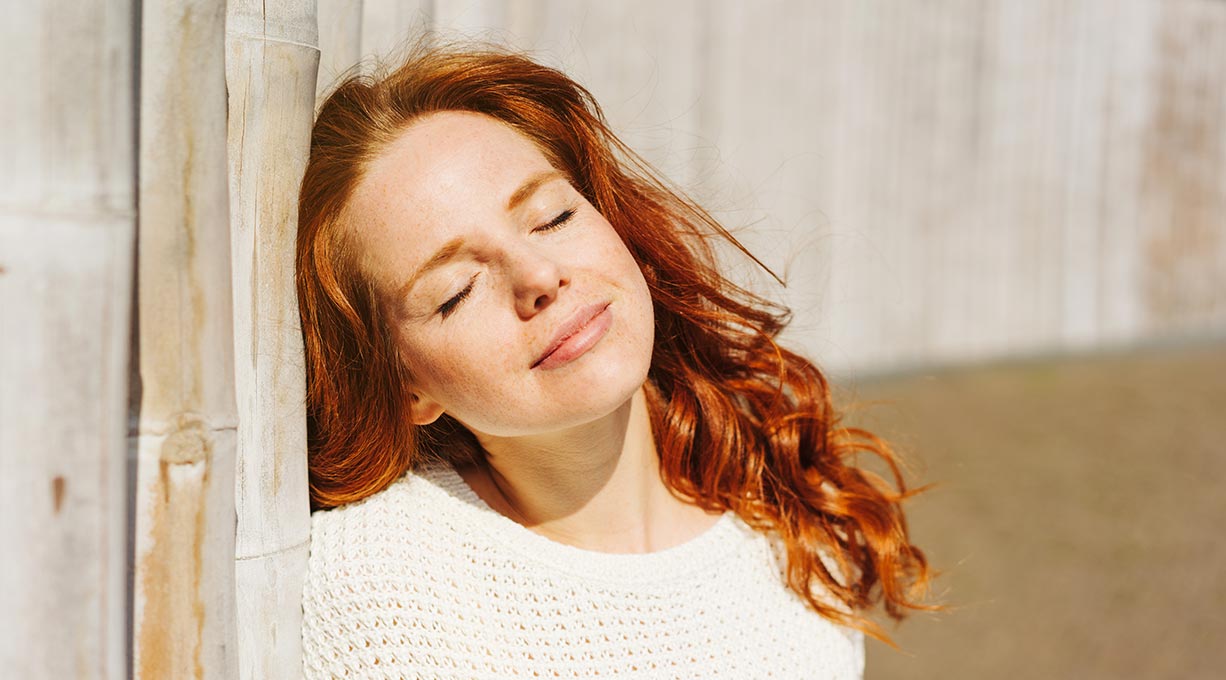 Frau mit roten Haaren lehnt an einer Wand und schließt die Augen in der Sonne