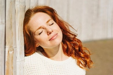 Frau mit roten Haaren lehnt an einer Wand und schließt die Augen in der Sonne