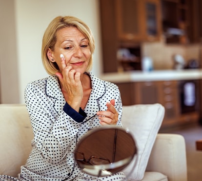 Frau im Pyjama trägt Gesichtscreme auf und betrachtet sich im Spiegel auf dem Sofa