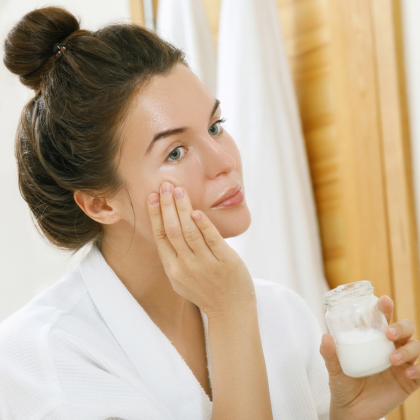 Frau im Bademantel trägt Kokosöl auf die Wange auf und hält ein Glas mit Kokosöl