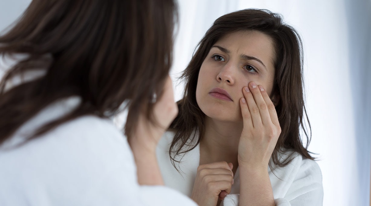 Frau mit braunen Haaren betrachtet sich skeptisch im Spiegel und berührt die Wange