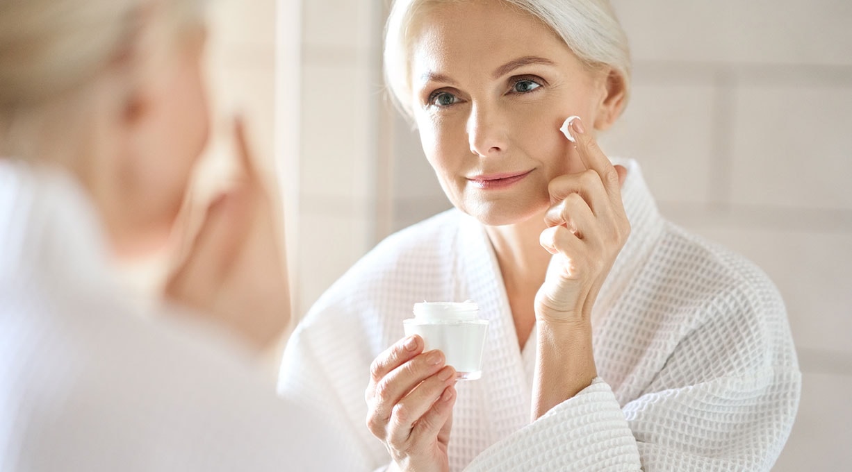 Eine Frau im Bademantel betrachtet sich lächelnd im Spiegel, während sie Creme aufträgt.