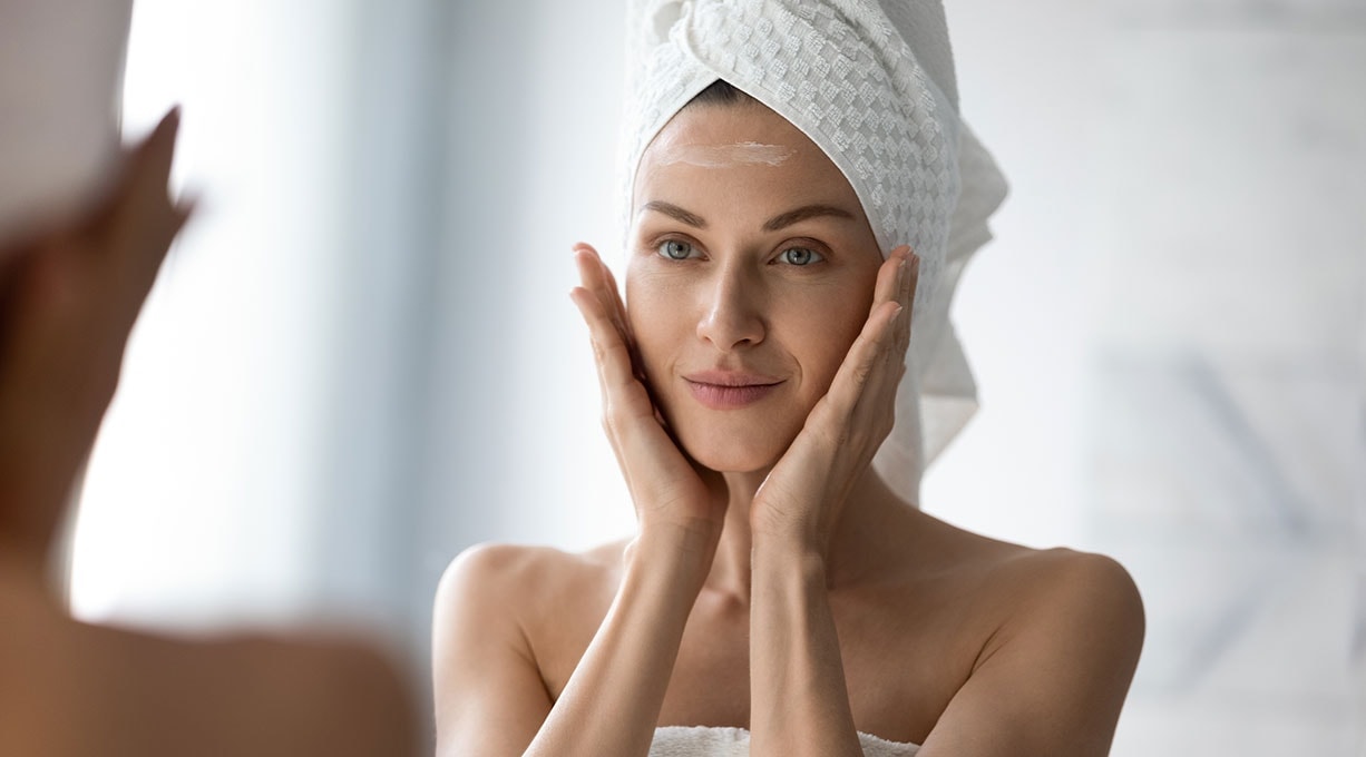 Frau mit Handtuch auf dem Kopf berührt die Wangen und trägt Gesichtscreme auf die Stirn auf