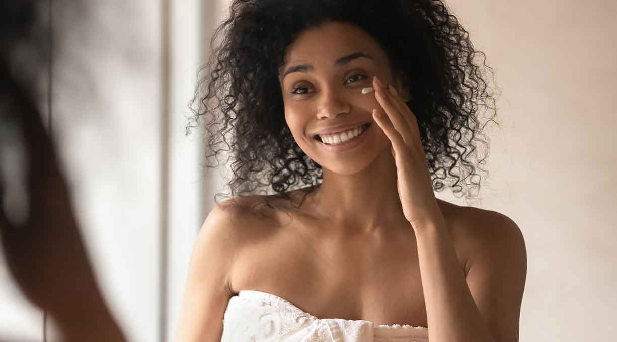 Eine lächelnde Frau betrachtet sich im Spiegel und trägt Creme auf ihr Gesicht auf.
