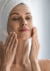 Eine Frau mit Handtuch auf dem Kopf cremt sich im Gesicht ein