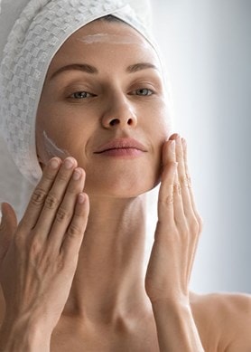 Eine Frau mit Handtuch auf dem Kopf cremt sich im Gesicht ein