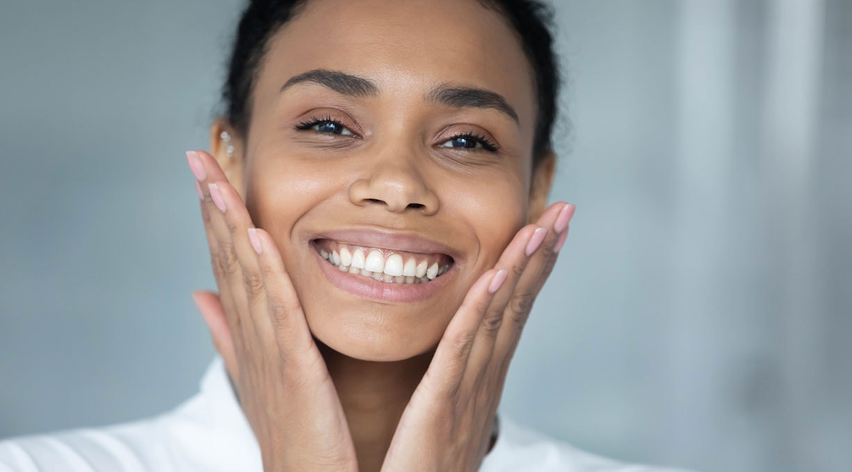 Lachende Frau berührt mit beiden Händen ihr Gesicht und zeigt gesunde Haut