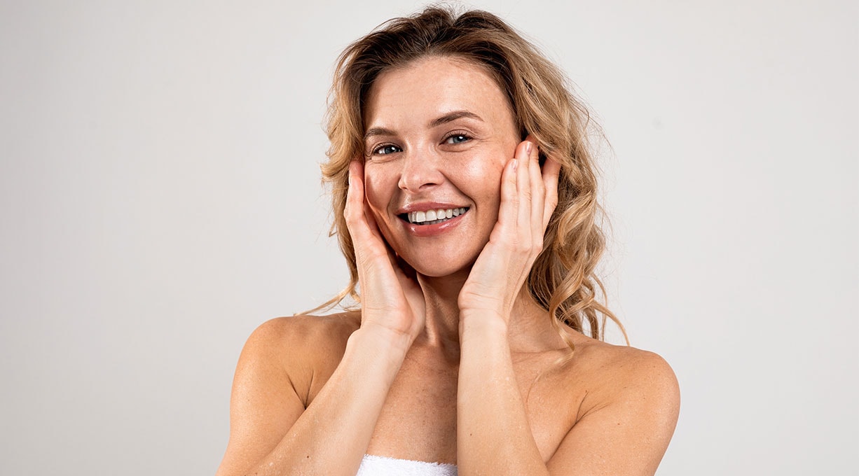 Lächelnde Frau mit Handtuch auf den Schultern berührt nach Hautpflege ihre Wangen