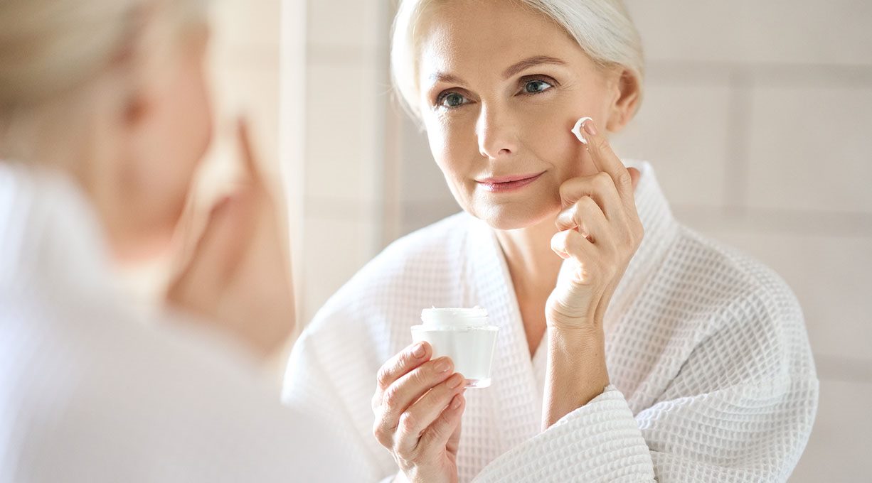 Ältere Frau trägt im Spiegel Gesichtscreme zur Anti-Aging-Pflege auf