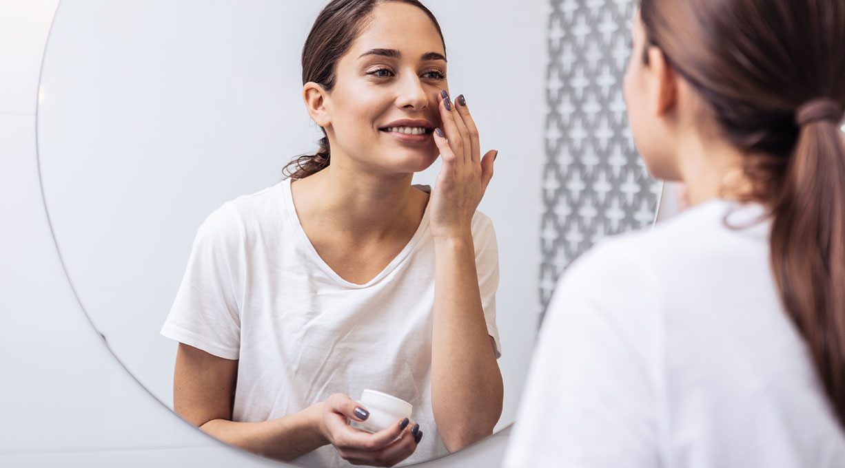 Eine Frau schaut in den Spiegel und lächelt, während sie Creme aufträgt. 