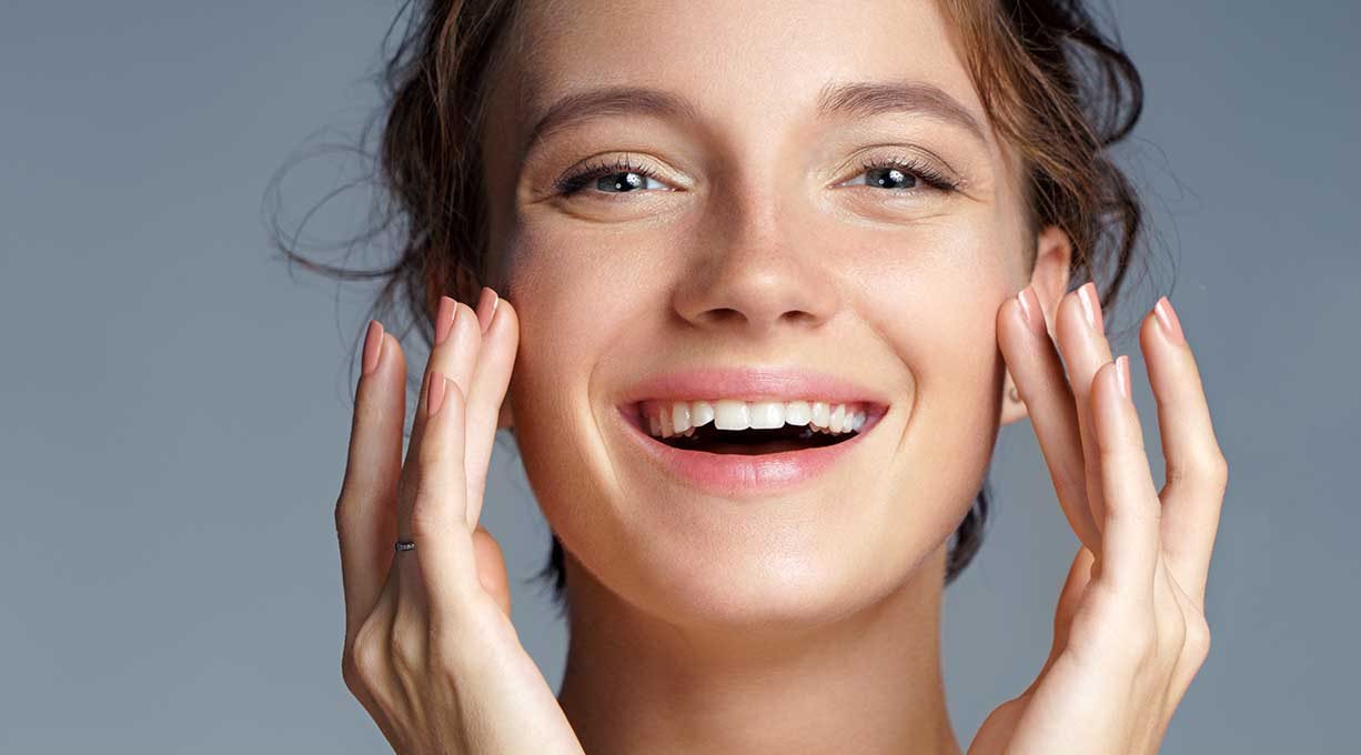 Eine Frau lächelt fröhlich und berührt mit den Fingern ihre Wangen. 