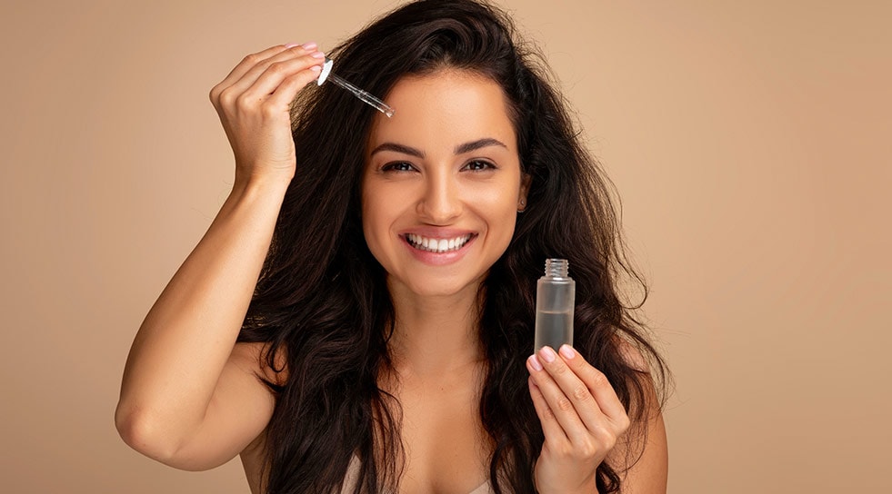 Frau mit langen Haaren appliziert Gesichtsserum mit Pipette und lächelt
