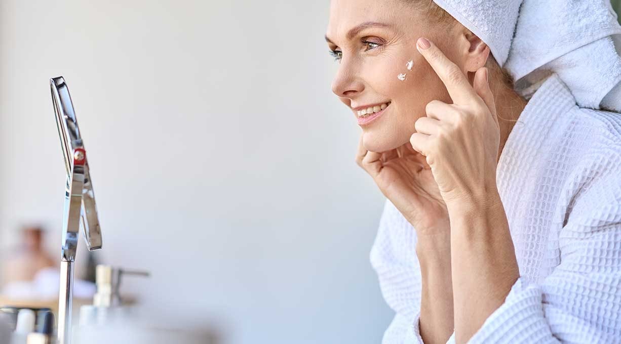 Stirnfalten glätten: Eine Frau mit Fältchen um die Augen tupft sich vor einem Spiegel Creme in ihr Gesicht