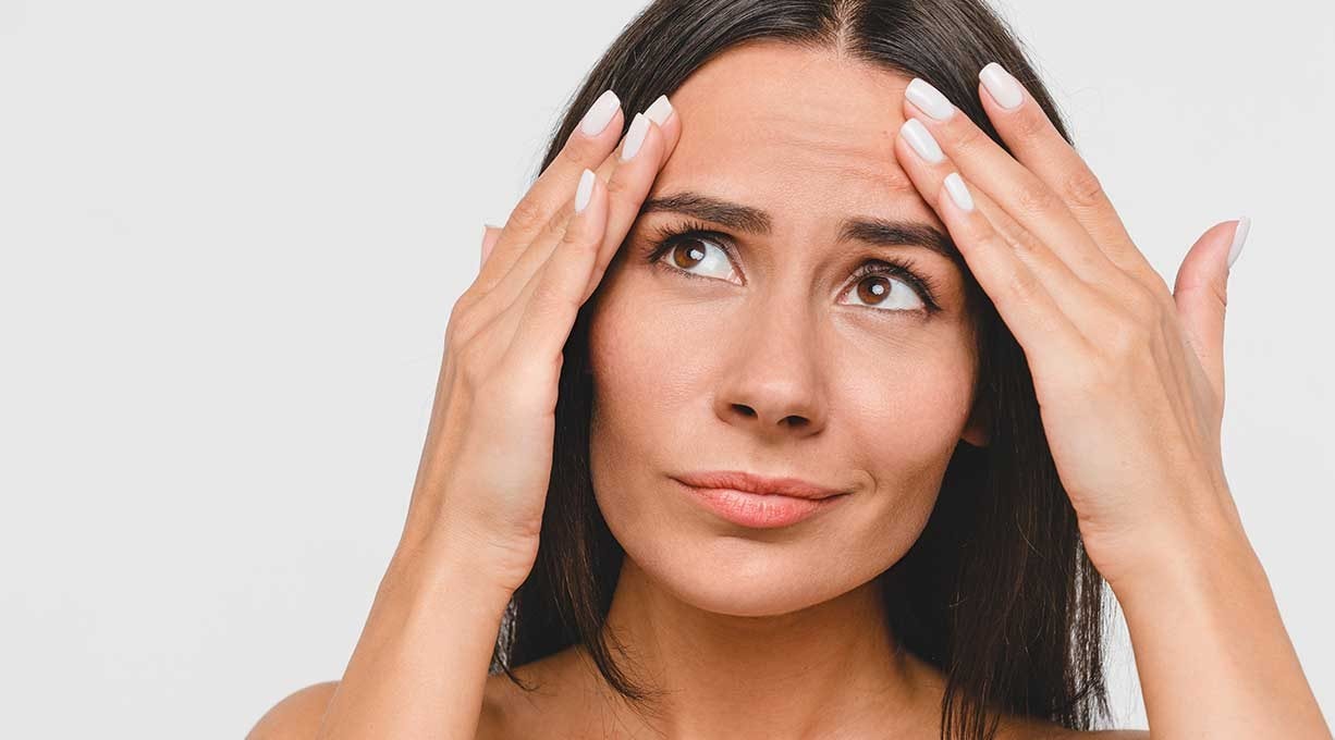 Eine Frau mit besorgtem Blick streicht mit den Fingern beider Hände über ihre Stirnfalten 