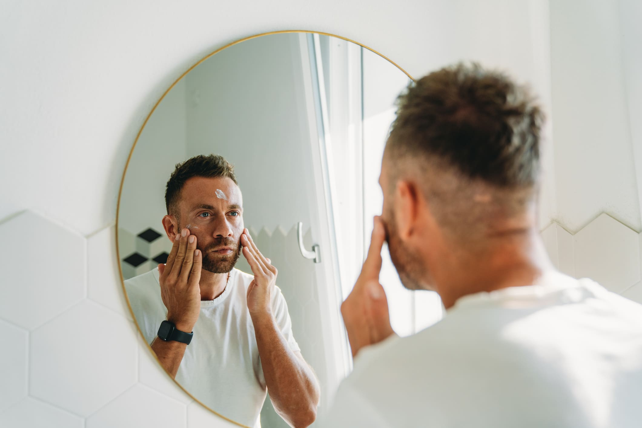 Ein Mann trägt im Spiegel Gesichtspflege auf, morgendliche Hautpflegeroutine.