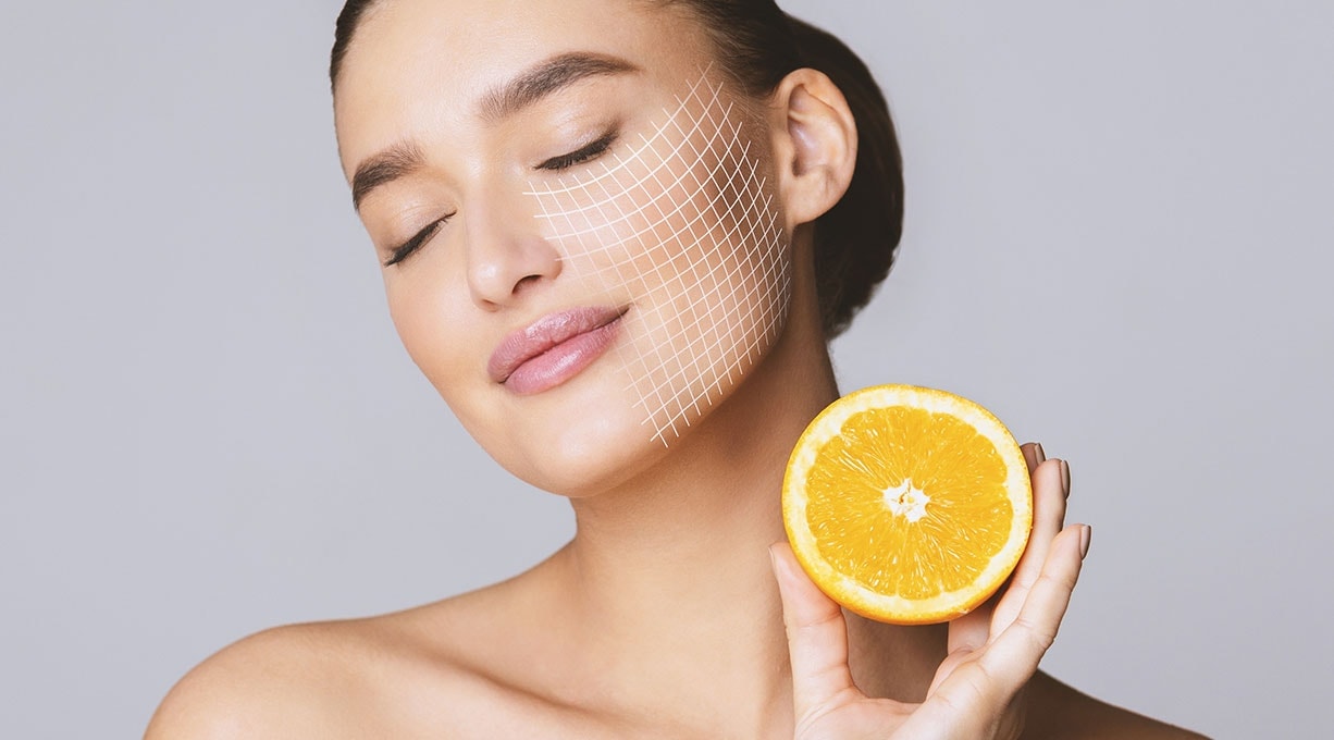 Frau hält halbe Orange neben Gesicht, Symbol für Vitamin C in Hautpflege.