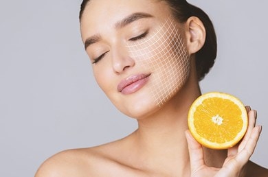 Frau hält halbe Orange neben Gesicht, Symbol für Vitamin C in Hautpflege.