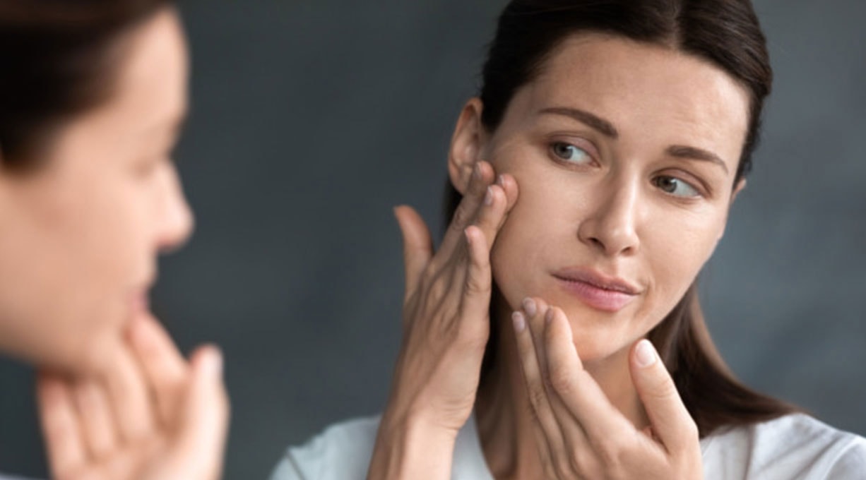 Frau prüft ihre Gesichtshaut im Spiegel, Fokus auf Hautelastizität und Pflege.