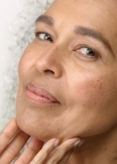 Reife Frau mit grauen Locken berührt sanft ihr Gesicht und zeigt gepflegte Haut.