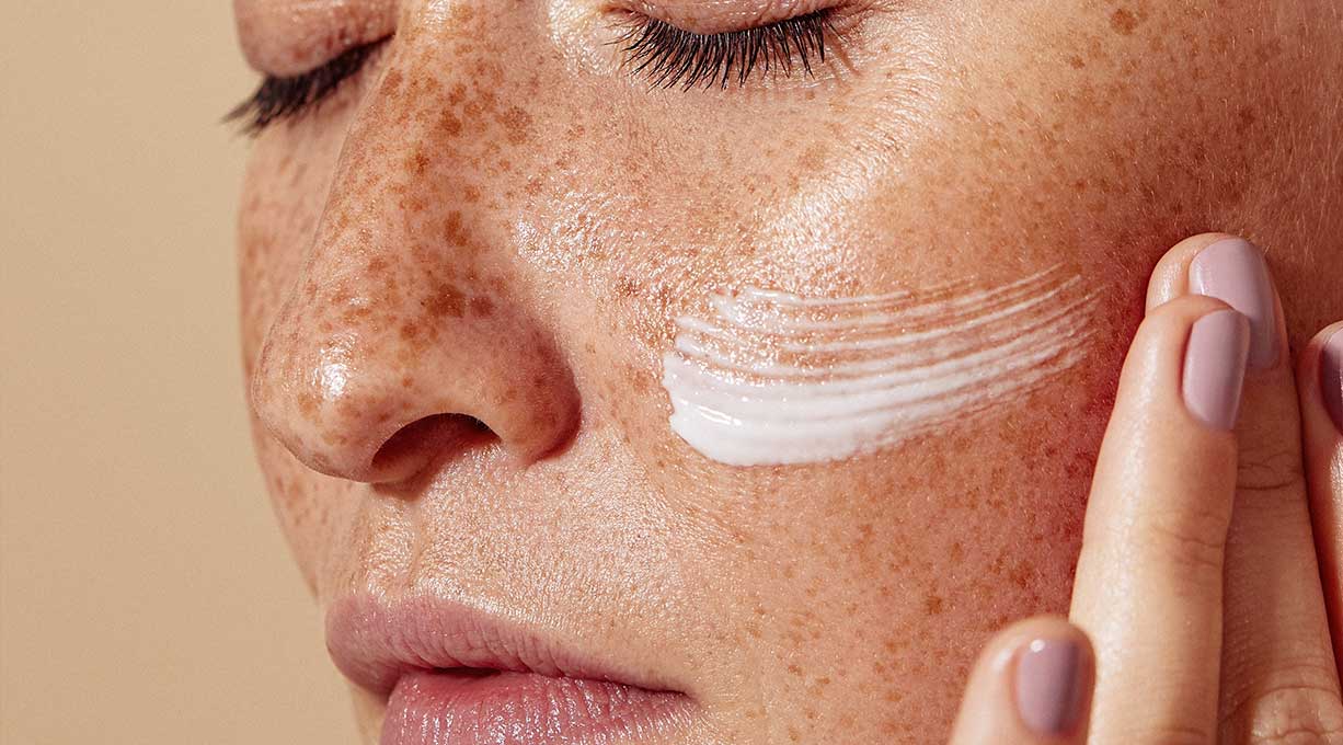 Frau trägt Gesichtscreme auf, Pflege für feuchtigkeitsarme Haut mit Sommersprossen