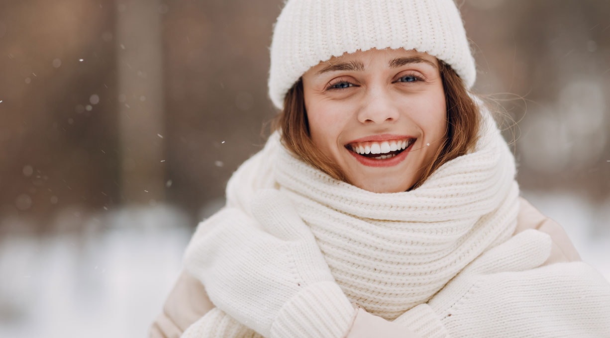 Eine lächelnde Frau mit weißer Mütze, weißem Schal und weißen Handschuhen in einer winterlichen Landschaft