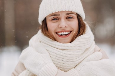 Eine lächelnde Frau mit weißer Mütze, weißem Schal und weißen Handschuhen in einer winterlichen Landschaft