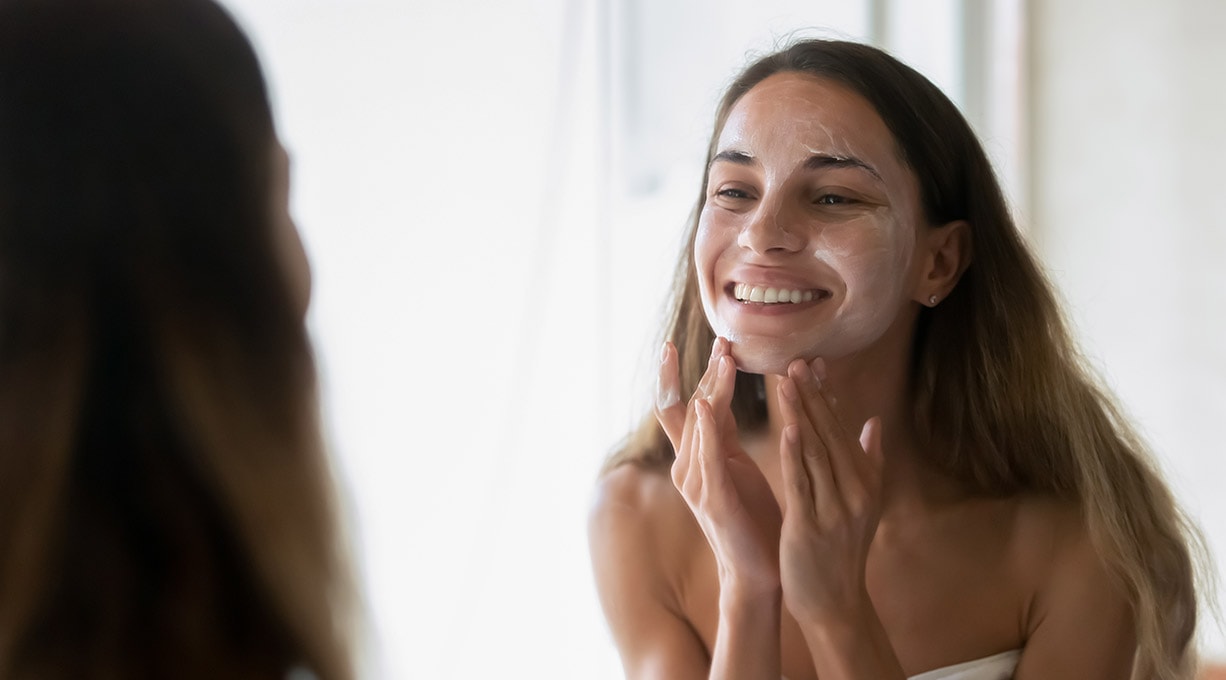 Eine Frau steht vor dem Spiegel und trägt Hautpflege im Winter auf ihr Gesicht auf