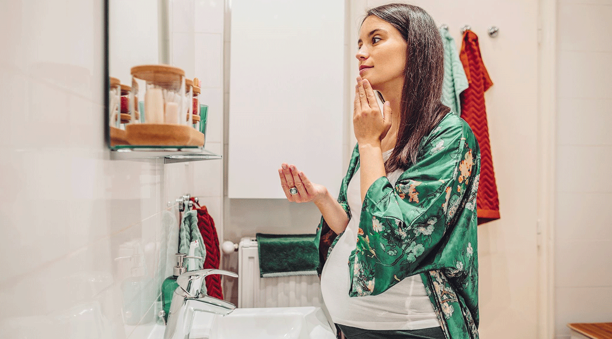 Schwangere Frau cremt ihr trockene Haut im Gesicht vor einem Spiegel im Bad ein