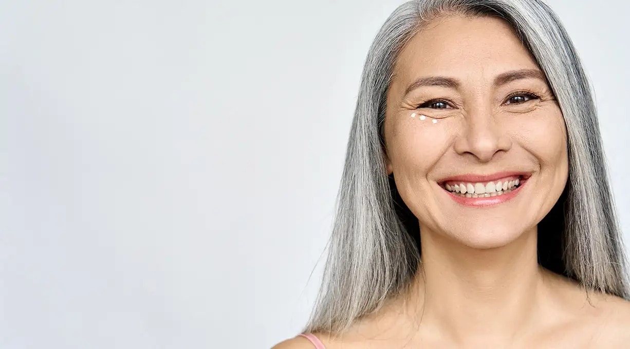 Ältere Frau mit grauen Haaren hat punktuell Augencreme aufgetragen und lächelt