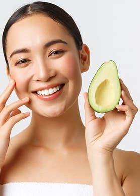 Junge Frau lächelt und berührt ihr Gesicht während sie eine Avocado in der Hand hält