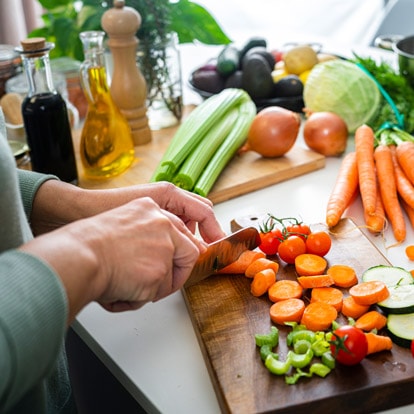 Hände schneiden Gemüse auf einem Schneidebrett in der Küche, umgeben von frischen Zutaten wie Karotten, Zucchini und Tomaten
