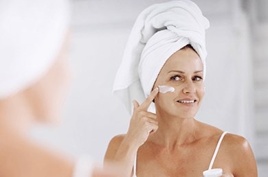 Frau mit Haarturban blickt in den Spiegel und cremt ihr Gesicht ein