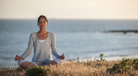 Eine Frau meditiert im Schneidersitz im Gras, im Hintergrund sieht man das Meer.