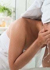 Frau mit Handtuch am Körper und auf dem Kopf gibt vor einem Waschbecken stehend Wasser ins Gesicht