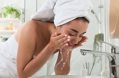 Frau mit Handtuch am Körper und auf dem Kopf gibt vor einem Waschbecken stehend Wasser ins Gesicht