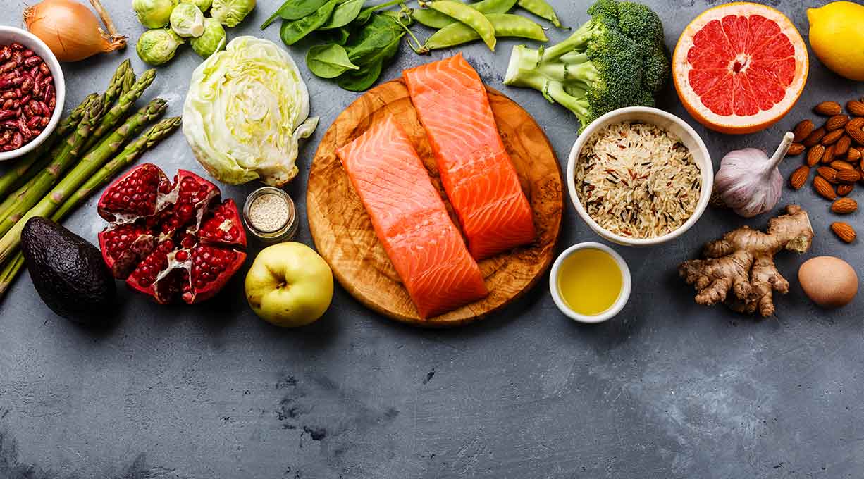  Frische gesunde Lebensmittel auf einem grauen Untergrund, darunter Lachsfilets auf einem Holzbrett, grünes Gemüse wie Brokkoli, Spinat und Spargel, Obst wie Granatapfel, Grapefruit und Apfel, Vollkornreis, Nüsse, Ingwerwurzel, Avocado, Knoblauch und Olivenöl.