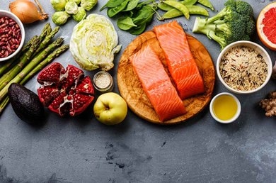 Frische gesunde Lebensmittel auf einem grauen Untergrund, darunter Lachsfilets auf einem Holzbrett, grünes Gemüse wie Brokkoli, Spinat und Spargel, Obst wie Granatapfel, Grapefruit und Apfel, Vollkornreis, Nüsse, Ingwerwurzel, Avocado, Knoblauch und Olivenöl.