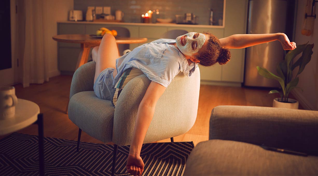 Junge Frau mit Gesichtsmaske im Gesicht streckt sich gemütlich auf einem Sessel aus