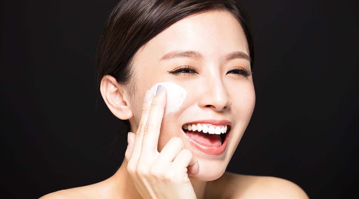 Eine fröhliche Frau trägt Glykolsäure-Creme auf ihre Wange auf 