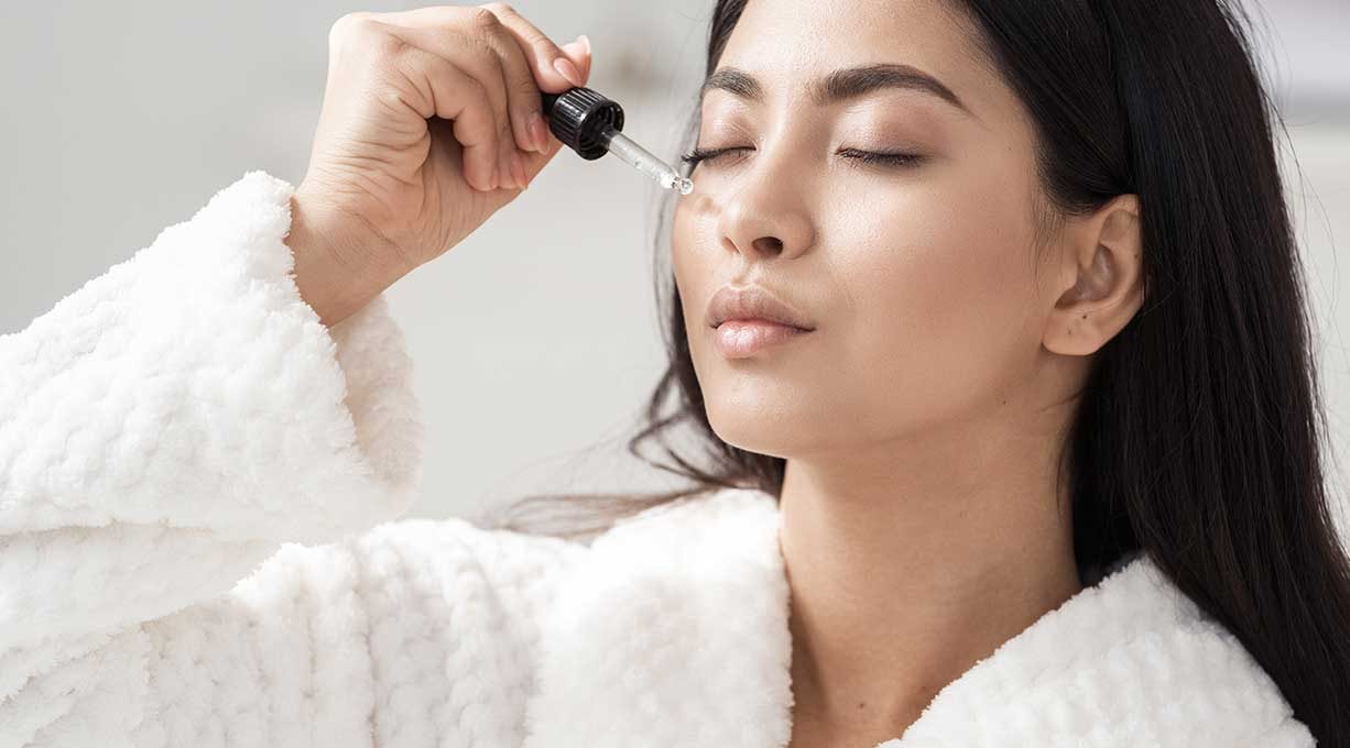 Eine Frau mit geschlossenen Augen gibt sich Serum mit einer Pipette auf die Wange 