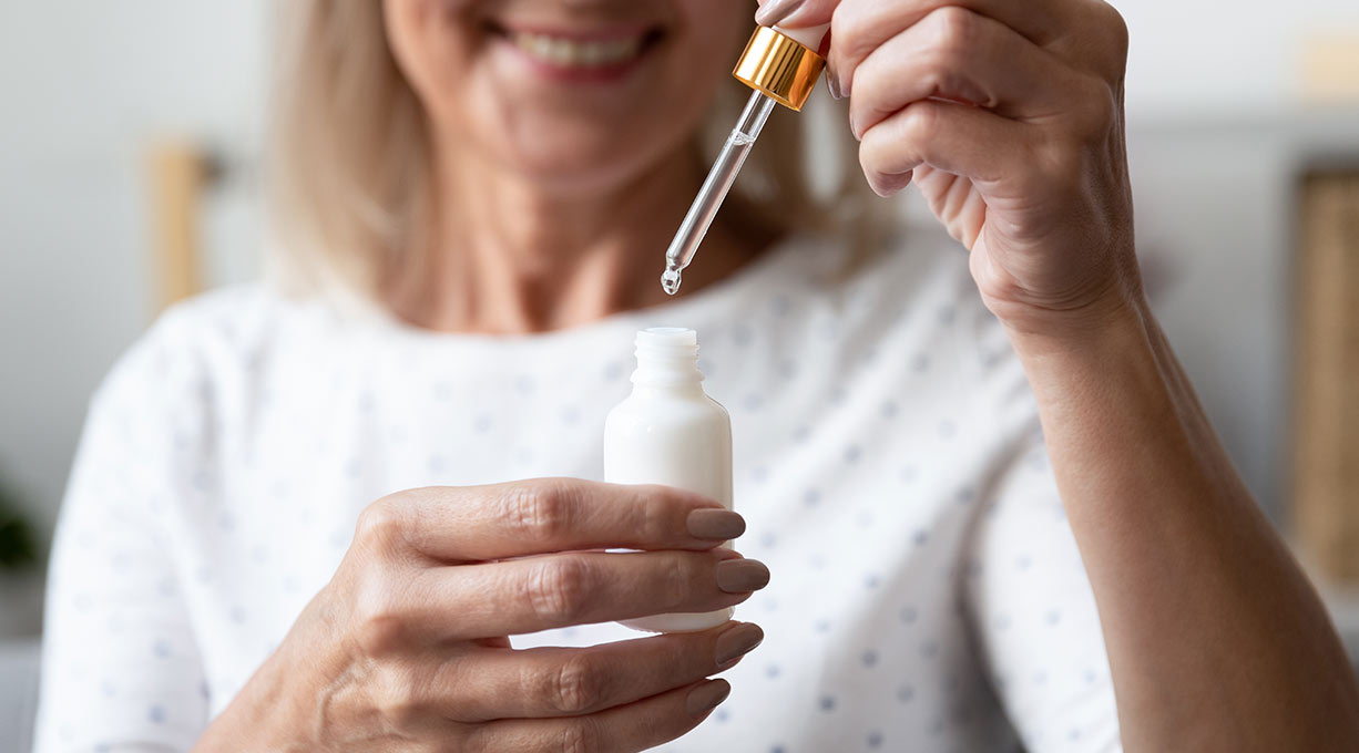 Ältere Frau lächelt während sie ein Gesichtsserum aus einer Pipettenflasche in der Hand hält